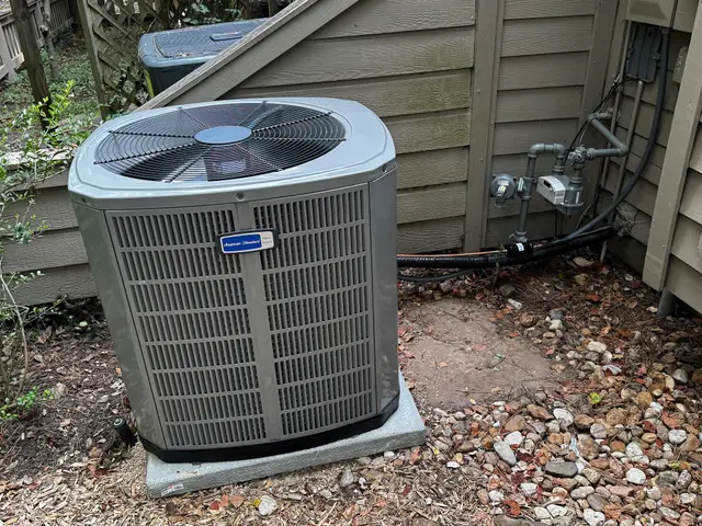 An American Standard condenser installed beside the siding of a home, gas meter and line set visible